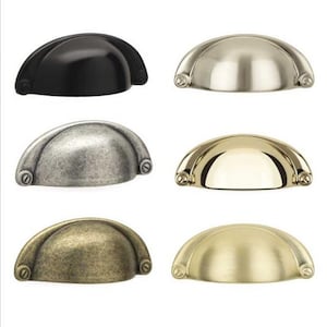 Könnte beinhalten: Eine Sammlung von sechs halbkreisförmigen Muschelgriffen für Schubladen, angeordnet in zwei Spalten. Die Oberflächen umfassen Mattschwarz, poliertes Nickel, antikes Silber, poliertes Messing, antikes Messing und gebürstetes Gold.
