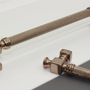 May include: Two brushed rose gold cabinet pulls with a square base and a textured, cylindrical handle.