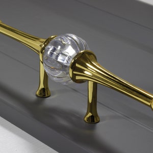 May include: A gold-toned cabinet pull with a clear, faceted glass sphere in the centre. The pull has a rounded, decorative end on each side.