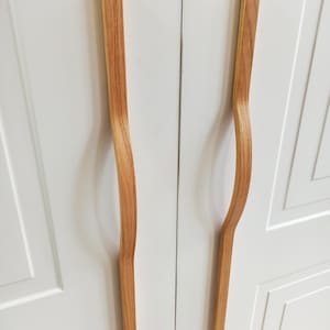 Puede incluir: Dos tiradores de armario de madera con acabado natural. Los tiradores tienen un diseño curvo y ergonómico y están montados en una puerta de armario blanca. La veta de la madera es visible, añadiendo calidez al diseño minimalista.