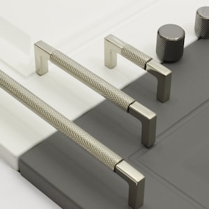 Può includere: Un set di quattro pomelli e maniglie per armadi argentati con un motivo a diamante testurizzato. I pomelli sono rotondi e le maniglie sono rettangolari con un angolo di 90 gradi.