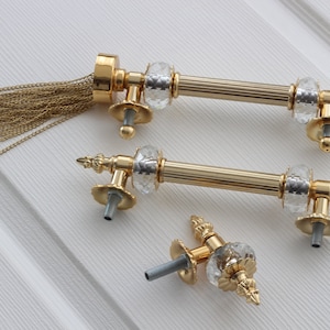Könnte beinhalten: Drei goldene Türklinken mit Kristall-Akzenten. Die Griffe haben eine dekorative Quastenkette.