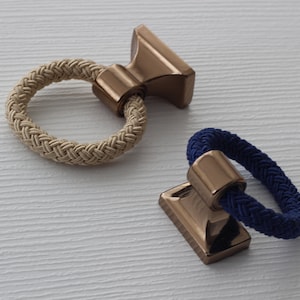 以下が含まれることがあります： 2つの装飾的なキャビネットプル。ベージュのロープループとネイビーブルーのロープループがあり、どちらもブロンズ色の長方形のベースに取り付けられています。白いテクスチャード加工の背景に配置されています。