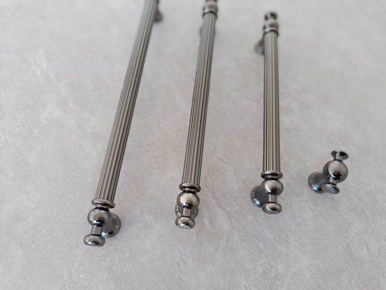 May include: Four silver cabinet pulls with a fluted design and round knobs. The pulls are arranged in a row on a light grey surface.