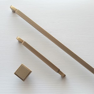 Puede incluir: Un juego de tres piezas de herrajes para gabinetes de color bronce. El juego incluye dos tiradores de barra rectangulares y un pomo cuadrado. Los tiradores tienen un diseño acanalado y el pomo un patrón cuadrado a juego. El hardware se muestra sobre una superficie blanca texturizada.