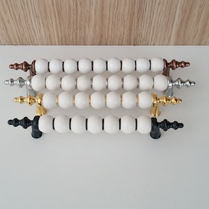 May include: Four cabinet pulls with white beaded handles. The pulls have different metal finishes: bronze, silver, gold, and black. The handles are arranged horizontally against a white surface.