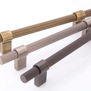 Op de afbeelding: Drie horizontale meubelgrepen met een geribbeld, cilindrisch ontwerp. De grepen zijn afgewerkt in metallic tinten van antiek messing, geborsteld nikkel en donkerbrons, elk voorzien van twee rechthoekige montagevoeten voor bevestiging.