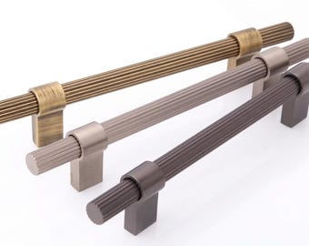 May include: Three horizontal cabinet pulls with a fluted, cylindrical design. The pulls are finished in metallic tones of antique brass, brushed nickel, and dark bronze, each featuring two rectangular mounting posts for attachment to furniture.