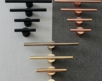 Può includere: Una collezione di maniglie per mobili moderne in varie finiture metalliche. Le maniglie sono disposte in file, mostrando tonalità nere, rame e oro. Ogni maniglia presenta un design a barra orizzontale, montata su una base circolare.