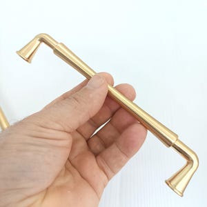 May include: A gold-colored cabinet pull with a straight bar and angled ends. The pull is held in a hand against a white background. The ends of the pull have a flared, conical shape.