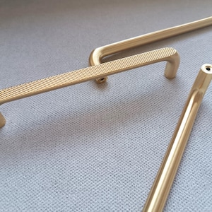 May include: Three gold-toned cabinet door pulls with a textured finish. Two of the pulls are short and have a curved shape, while the third pull is long and straight.