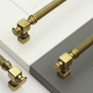 Può includere: Primo piano di maniglie per mobili in ottone antico. Le maniglie hanno un motivo a diamante sulla barra e una base quadrata con una parte superiore rotonda. Le maniglie sono su ante di armadi bianche e grigie.