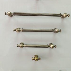 Puede incluir: Tres tiradores de puerta de armario de color dorado antiguo con un diseño decorativo. Los tiradores están dispuestos verticalmente, con el más grande en la parte superior y el más pequeño en la parte inferior. El tirador más pequeño es un simple pomo.