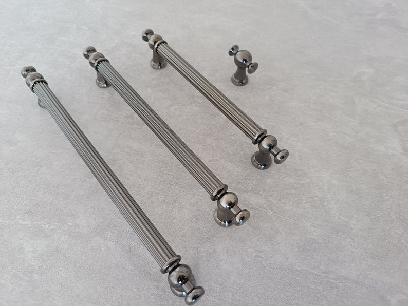 May include: Three black metal cabinet pulls with a fluted design and round knobs. The pulls are arranged in a row from longest to shortest.