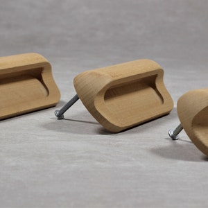 Könnte beinhalten: Drei Holzknaufgriffe mit rechteckigem Ausschnitt. Die Griffe sind hellbraun und haben eine leicht geschwungene Form. Jeder Griff hat eine silberne Schraube.