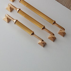 Op de afbeelding: Drie gouden kastgrepen met een geribbeld ontwerp. De grepen zijn van verschillende grootte en zijn gerangschikt in een rij van groot naar klein.