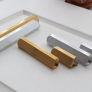 Può includere: Una collezione di maniglie per porte moderne in finitura oro e argento. Le maniglie hanno una forma cilindrica con una texture scanalata. L'hardware è esposto su una superficie bianca, mostrando il design e la finitura delle maniglie.