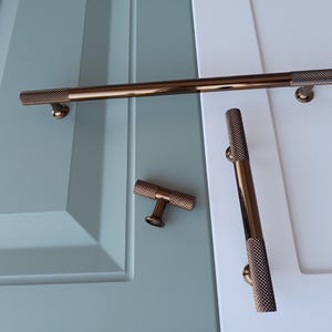 May include: Three bronze cabinet pulls with a textured finish. The pulls are in different sizes and shapes. The largest pull is a long bar with a rounded end. The medium pull is a shorter bar with a rounded end. The smallest pull is a small knob with a rounded end.