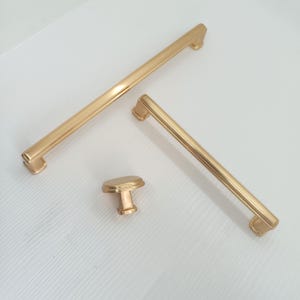 May include: Three gold cabinet hardware pulls. Two are long, rectangular pulls with rounded edges. One is a smaller, round knob with a square base.
