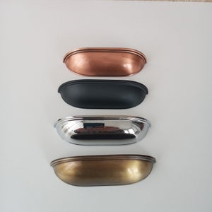 May include: Four cabinet pulls in different finishes: copper, black, chrome, and antique brass. Each pull has a rounded, oval shape with a raised edge.