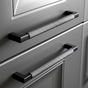 Op de afbeelding: Close-up van twee zwarte kastgrepen met een getextureerde, zilverkleurige grip. De handgrepen zijn rechthoekig met een haakse bocht aan elk uiteinde. Ze zijn gemonteerd op een grijze kastdeur.