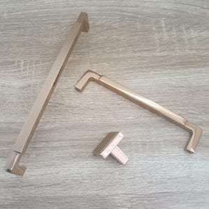 Op de afbeelding: Drie goudkleurige kastbeslagknoppen. De grootste knop is een lange, rechthoekige staaf met een getextureerd oppervlak. De middelgrote knop is een gebogen staaf met een glad oppervlak. De kleinste knop is een vierkante knop met een glad oppervlak.