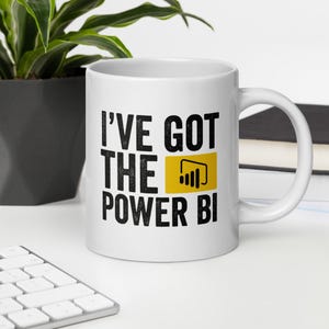 Op de afbeelding: Witte keramische mok met een zwarte en gele afbeelding met de tekst "I've got the Power BI".