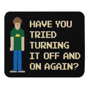 Op de afbeelding: Een zwarte muismat met een gepixelde afbeelding van een persoon met bruin haar en een groen shirt met een gele alien-afbeelding. De tekst "HAVE YOU TRIED TURNING IT OFF AND ON AGAIN?" staat in witte gepixelde letters.