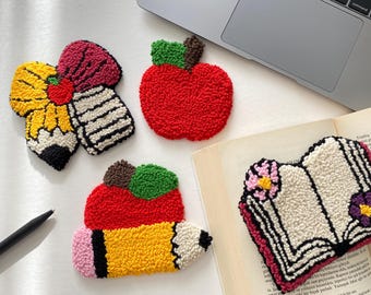 May include: A collection of colorful, textured, handmade coasters in the shape of school-themed items. The set includes a red apple, a pencil, a book, and a pencil with a flower.