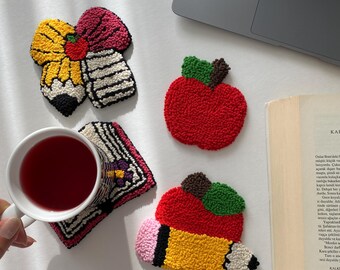 May include: A collection of colorful, handmade coasters in various shapes, including a red apple, a yellow pencil, and a pencil and eraser design. The coasters are placed on a white surface, with a cup of tea and a laptop in the background.