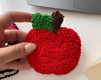 May include: A handmade, red apple-shaped rug with green and brown accents. The rug is made with a textured, looped yarn technique. The apple is held by a hand, with a laptop in the background.
