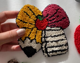 May include: A colorful, handmade textile art piece resembling a cupcake. The design features yellow, red, and white sections with black outlines. The cupcake has a red cherry on top and is held by a hand.