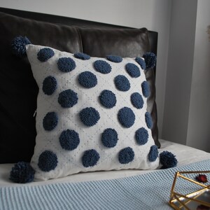 May include: A white throw pillow with blue pom-poms and blue embroidered circles. The pillow is on a blue bedspread with a gold decorative stand.