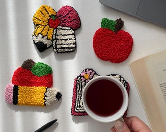 May include: A collection of colorful, handmade coasters in various shapes, including a pencil, apple, and floral designs. The coasters are made with a textured, looped yarn technique, and a cup of tea rests on one.
