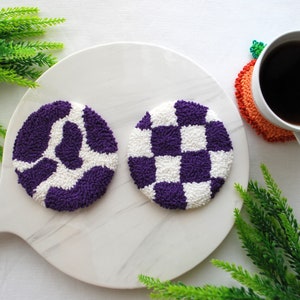 Könnte beinhalten: Zwei runde Untersetzer mit violetten und weißen Mustern auf einem weißen Marmor-Schneidebrett. Die Untersetzer sind mit einer Punch-Needle-Technik hergestellt.