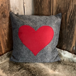 Puede incluir: Una almohada gris con un gran corazón rojo cosido en la parte delantera. La almohada está sobre una alfombra de piel sintética.