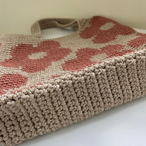 May include: A beige and dusty rose crochet tote bag with a floral pattern. The bag features a sturdy, textured design with a rectangular base and two handles. The flowers are a muted rose color against a beige background.