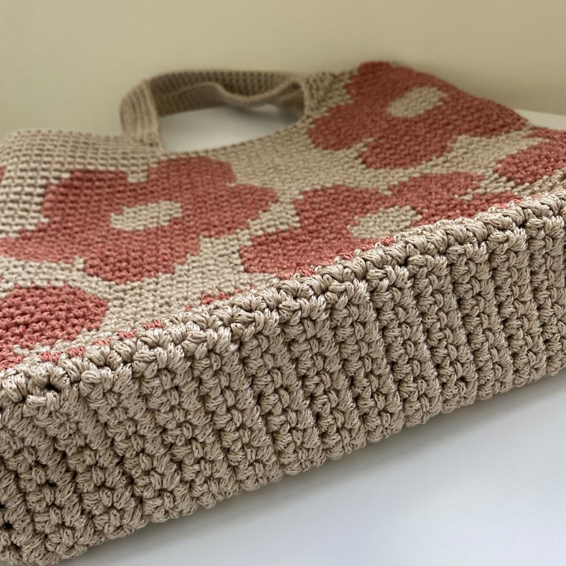 May include: A beige and dusty rose crochet tote bag with a floral pattern. The bag features a sturdy, textured design with a rectangular base and two handles. The flowers are a muted rose color against a beige background.