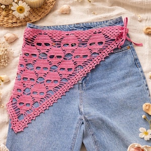 May include: A pink crochet skull bandana is draped over blue denim jeans. The triangular bandana features a repeating skull pattern. It is tied on the side with a pink string. The jeans are a light blue wash.
