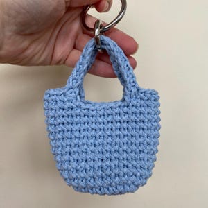 Puede incluir: Un llavero de mini bolso de crochet azul claro con un anillo de metal plateado.