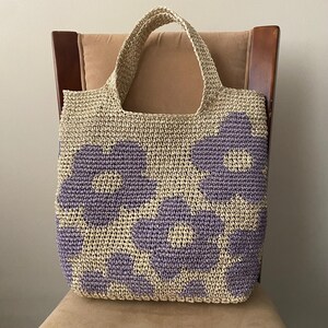 May include: A beige crocheted tote bag with large purple flower designs. The bag has two handles and is sitting on a chair. The bag is made of a woven material.
