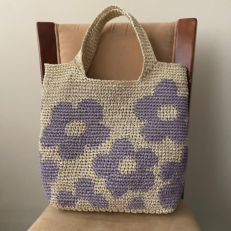 May include: A beige crocheted tote bag with large purple flower designs. The bag has two handles and is sitting on a chair. The bag is made of a woven material.