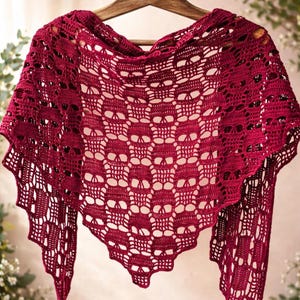 Pode incluir: Um xale triangular de crochê em vermelho escuro com um padrão intrincado e repetitivo de caveiras por todo o tecido. A peça é confeccionada com um ponto aberto delicado, criando uma aparência texturizada semelhante a renda com bordas festonadas.