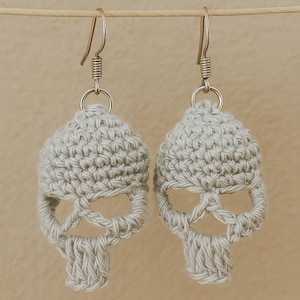 May include: A pair of light gray crochet skull earrings. The earrings feature a skull design with visible eye sockets, a nose cavity, and a jawline. The earrings are suspended from silver-colored ear wires.