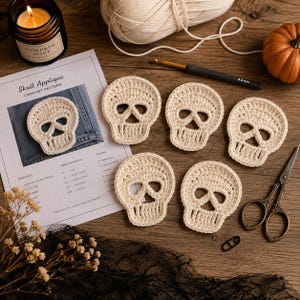 Puede incluir: Cinco apliques de calavera de ganchillo en color blanco roto dispuestos junto a una hoja de patrón de ganchillo impresa titulada Skull Applique, un ovillo de lana, un ganchillo de 4,5 mm, una calabaza pequeña, una vela, tijeras y un marcador.