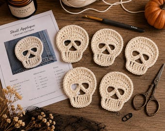 Peut inclure: Cinq appliques en forme de crâne crochetées en blanc cassé disposées à côté d'une fiche de modèle de crochet intitulée Skull Applique, un crochet de 4,5 mm, une pelote de laine, une petite citrouille, une bougie et des ciseaux en métal.