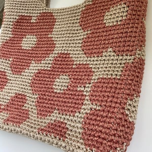 May include: A handmade crochet tote bag featuring a floral pattern. The bag is crafted with a combination of dusty rose and beige yarn, creating a textured design. The bag has a sturdy handle for carrying.