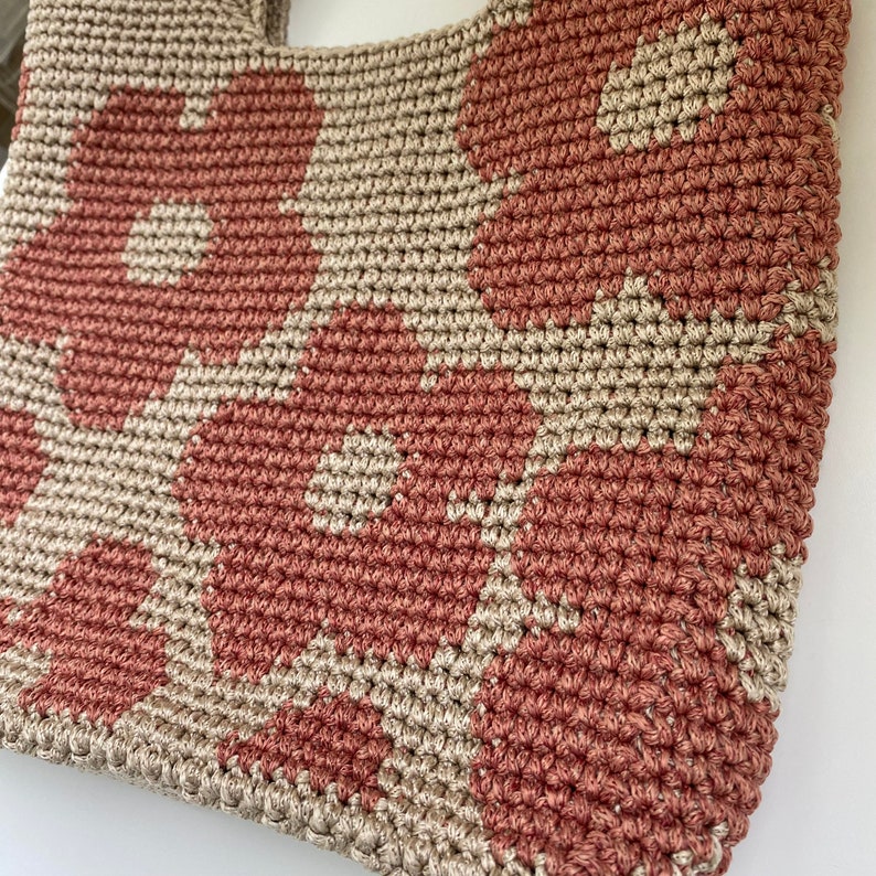 May include: A handmade crochet tote bag featuring a floral pattern. The bag is crafted with a combination of dusty rose and beige yarn, creating a textured design. The bag has a sturdy handle for carrying.