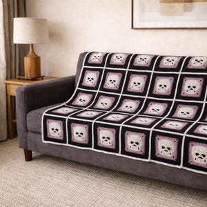 May include: A crocheted blanket with a repeating skull pattern in black, white, and mauve squares. The blanket is draped over a gray sofa. The skulls have a simple, stylized design. The blanket is a decorative home textile.