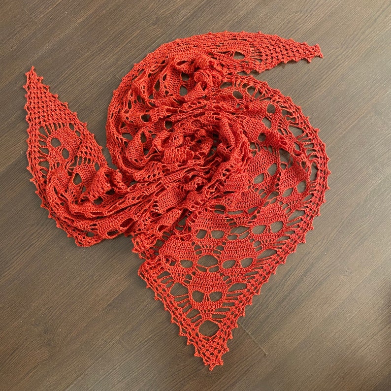Puede incluir: Un chal de ganchillo de color rojo oscuro con un delicado patr&oacute;n de encaje. El chal est&aacute; doblado y dispuesto en forma de espiral.